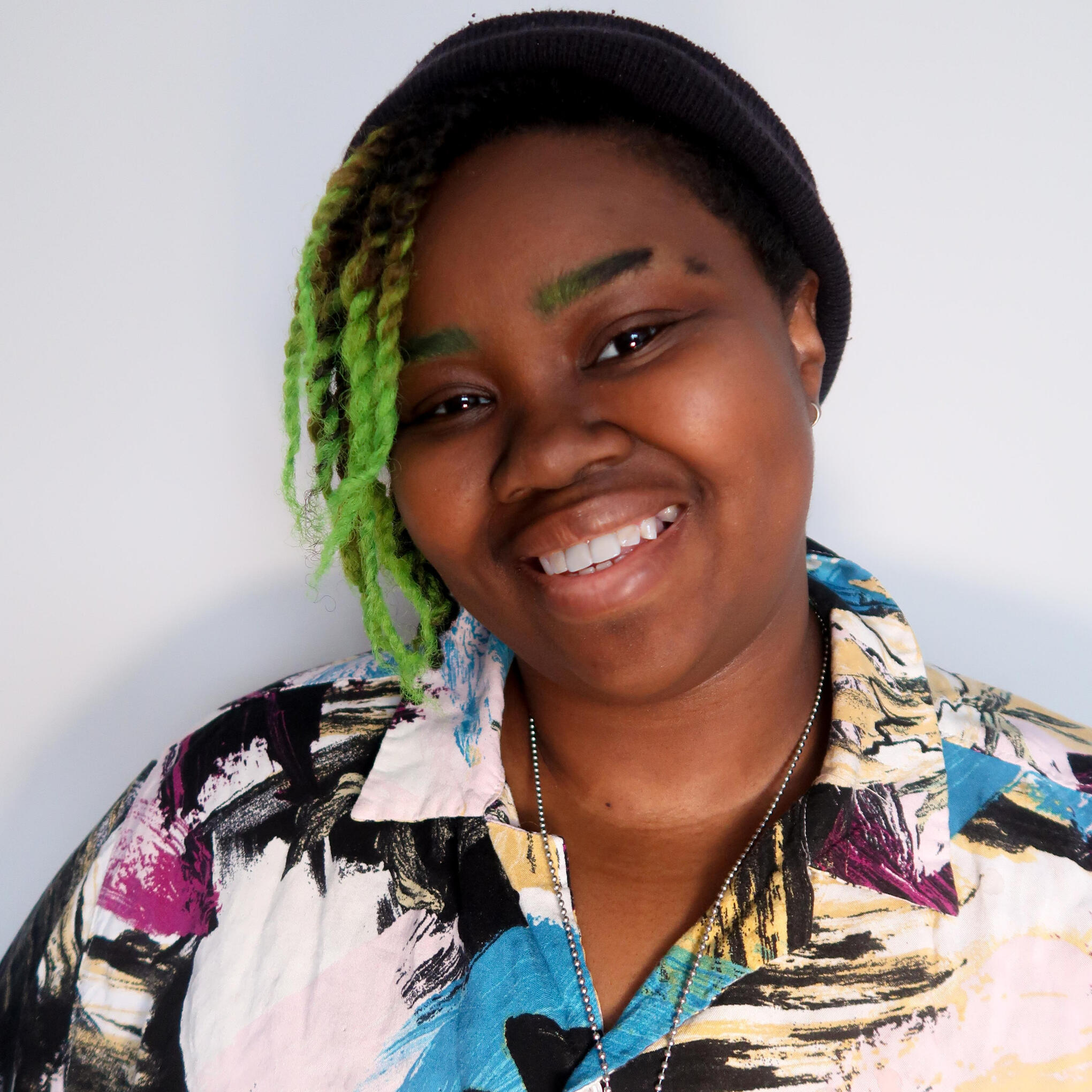 Dann R. Lynch A photo of Dann Lynch. A non-binary dark skin Afro-Indigenous person with hair in twists from black to bright green ombre on one side of their face their eyebrows match the ombre color and one has a deliberate slit in it. They are wearing a light, multicol